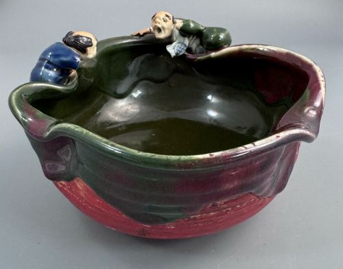 fig 3. A Japanese Sumida Gawa pottery bowl featuring a dark green running glaze over a red base with two sculpted figures leaning over the rim and an applied white makers mark.