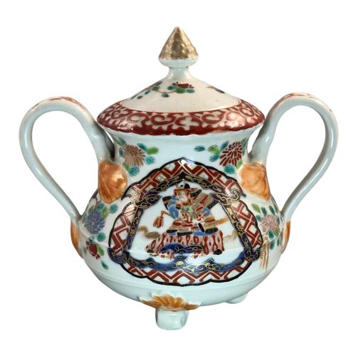 fig 3. A covered Japanese Imari porcelain sugar bowl featuring double side handles, a pointed gilded finial, and hand-painted samurai and floral motifs in red, blue, green, and gold enamels.