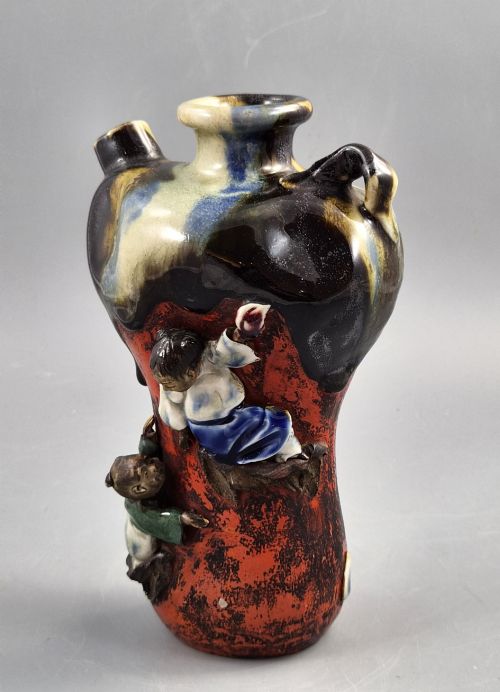 fig 1. A Japanese Sumida Gawa pottery vase featuring an asymmetrical spouted form with a dark flambe drip glaze over a textured red lower body adorned with applied high-relief figures of boys.