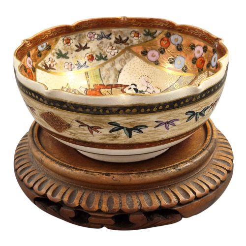 fig 2. A small lobed Japanese Satsuma ceramic bowl with intricate polychrome floral painting and gold gilding, resting on a carved dark wooden base.