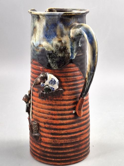 fig 1. A tall Japanese Sumida Gawa ceramic pitcher featuring a ribbed terracotta body with relief figures pouring sake and a dark drip glaze near the lip.