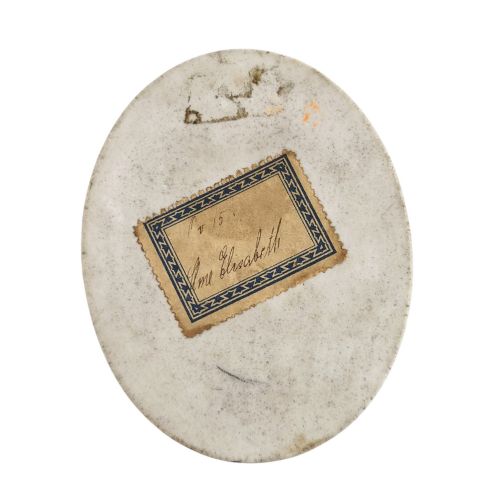 fig 3. An oval porcelain plaque featuring a hand-painted portrait of a woman with powdered hair wearing a purple dress and yellow shawl with a rose detail.