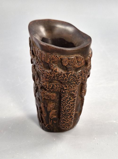 fig 1. A dark brown carved water buffalo horn libation cup featuring relief carvings of immortals standing under pine trees.