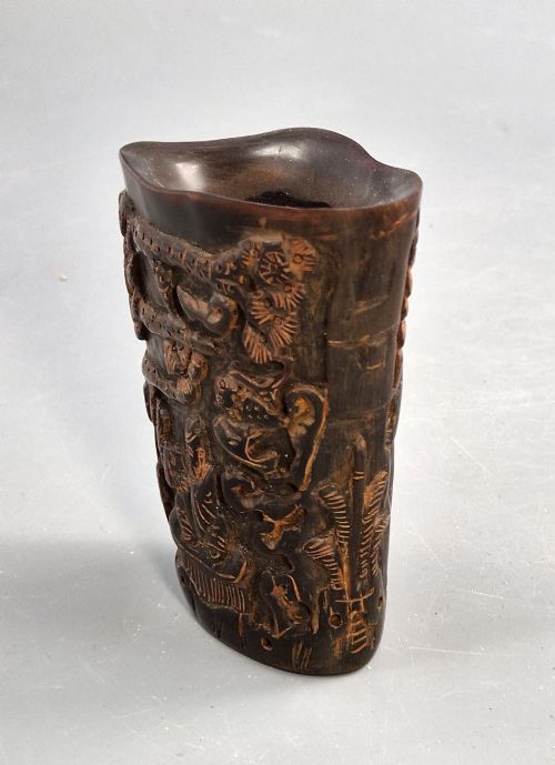fig 4. A dark brown carved water buffalo horn libation cup featuring relief carvings of immortals standing under pine trees.
