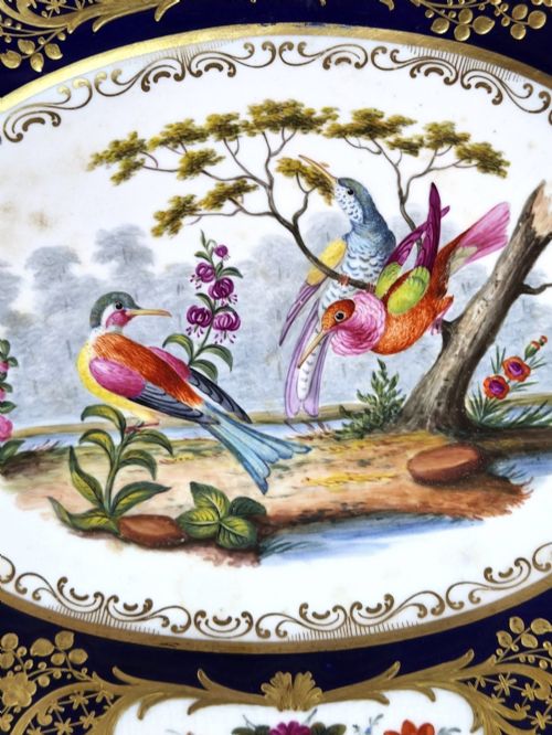 fig 1. An oval porcelain dish featuring a dark cobalt blue rim with gold scrollwork, reticulated handles, floral panels, and a central hand-painted scene of colorful birds perched on branches.