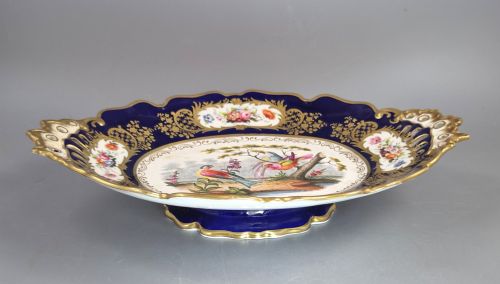 fig 4. An oval porcelain dish featuring a dark cobalt blue rim with gold scrollwork, reticulated handles, floral panels, and a central hand-painted scene of colorful birds perched on branches.