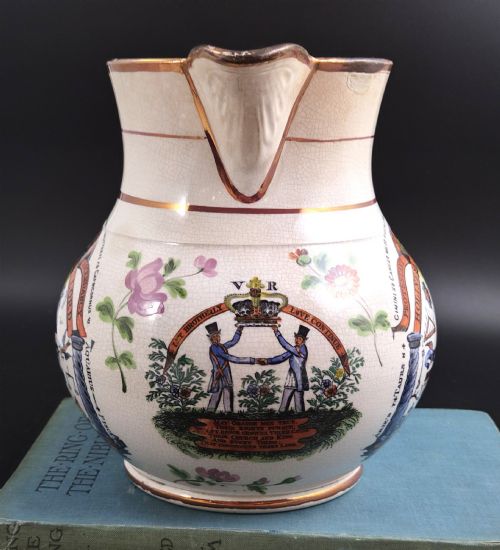 fig 1. A cream earthenware jug featuring hand-painted floral motifs, copper lustre trim, and a printed Orange Order emblematic coat of arms with religious and fraternal symbols.