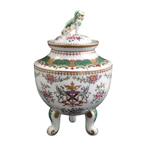 fig 1. A white hard-paste porcelain covered jar on three cabriole legs, decorated with floral enamel sprays, an armorial crest, green lattice borders, and topped with a foo dog lid finial.