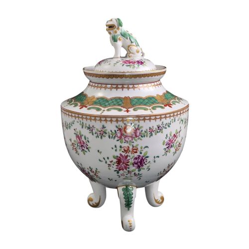 fig 3. A white hard-paste porcelain covered jar on three cabriole legs, decorated with floral enamel sprays, an armorial crest, green lattice borders, and topped with a foo dog lid finial.