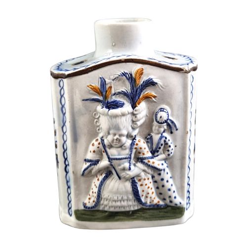 fig 1. An 18th-century Prattware earthenware tea canister featuring relief-moulded depictions of two Georgian Macaroni figures in blue, ochre, and green underglaze glazes against an off-white background.