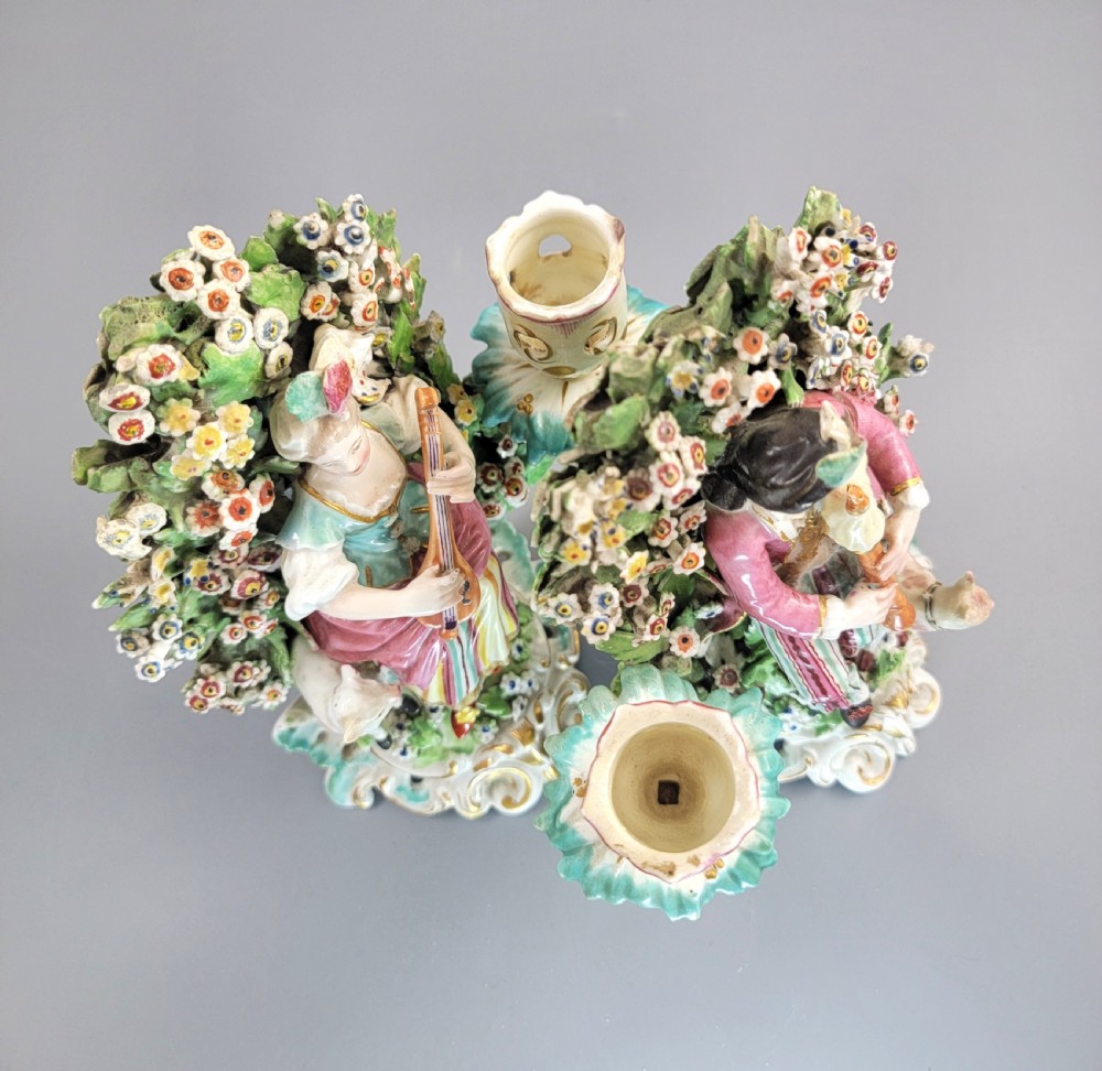 A pair of 18th-century soft-paste Derby porcelain candlesticks depicting a male and female musician seated before leafy floral bocage backdrops. View VII
