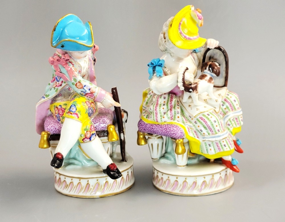 A pair of 18th-century hand-painted Meissen porcelain figurines depicting a boy and girl seated on stools with small dogs on circular fluted bases. View V