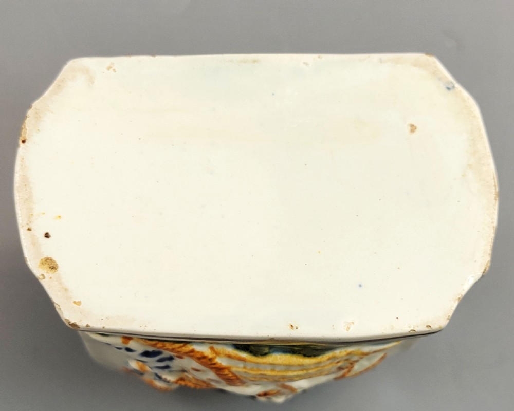 A late 18th-century cream-coloured pearlware tea caddy featuring moulded relief figures painted in ochre and cobalt blue glazes. View X