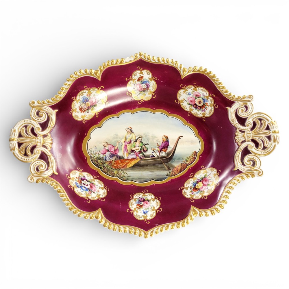 An oval early 19th-century claret porcelain serving dish featuring gold scroll handles, floral border panels, and a central hand-painted scene of figures in a boat.