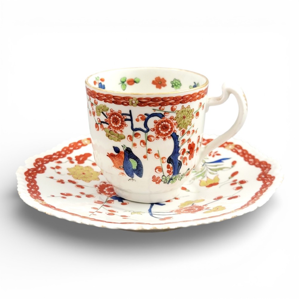 A white porcelain teacup and matching saucer adorned with hand-painted quail birds, red and green floral foliage, and gilded rims.