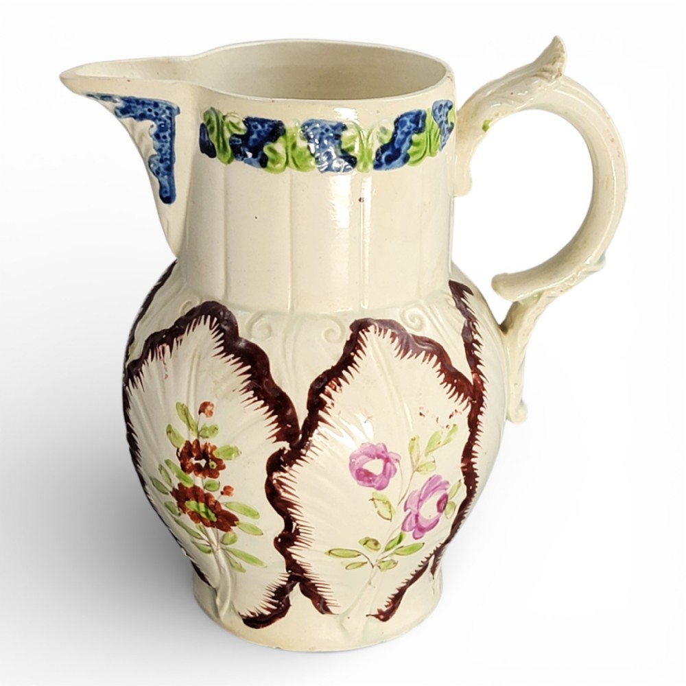 An antique cream glazed ceramic jug featuring raised cabbage leaf moulding, hand-painted floral motifs, and a moulded scroll handle.