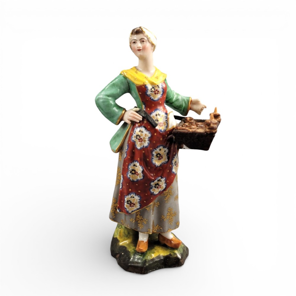 An antique Georgian Derby porcelain figurine depicting a female street vendor wearing a green jacket and patterned apron while holding a tray of baked goods.