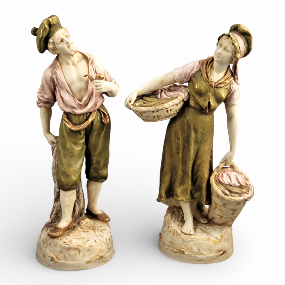 A pair of antique Royal Dux porcelain figures depicting a fisherman with a pipe and a fisherwoman carrying baskets of fish, finished in muted green, pink, and gilt glazes.