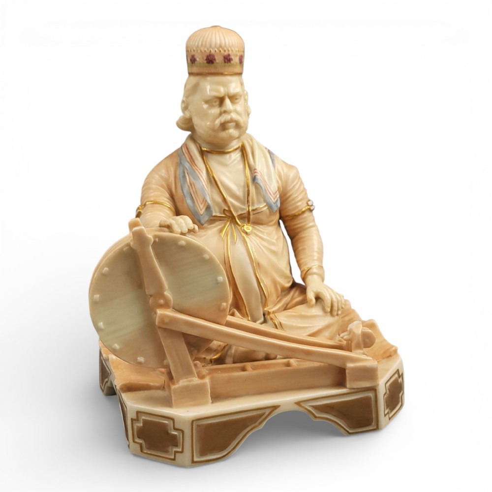 A late 19th-century Royal Worcester blush ivory porcelain figurine depicting an Indian craftsman seated at a spinning wheel, detailed with polychrome enamel and gold gilded highlights.