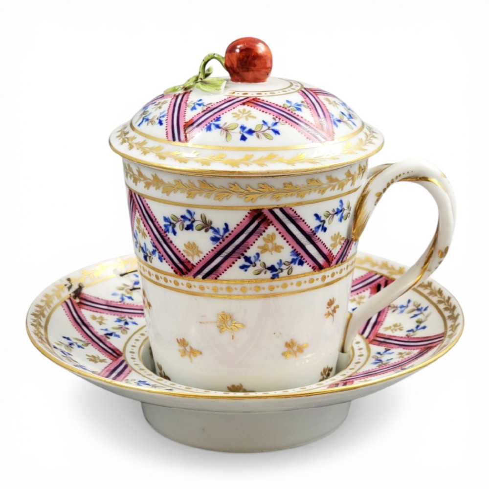 A French Neoclassical porcelain covered trembleuse cup and saucer decorated with pink ribbons, blue floral garlands, gold leaf borders, and a cherry-shaped lid finial.