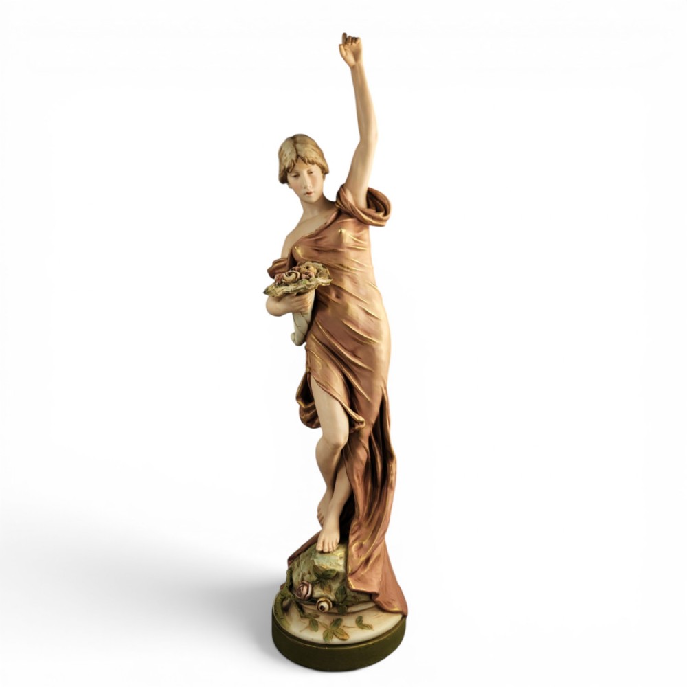 An Art Nouveau porcelain figure depicting a maiden in flowing drapery with one arm raised aloft and holding flowers at her waist.