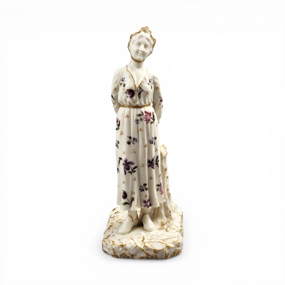 A standing porcelain figurine depicting a lady in a floral dress and bonnet with gilded trim on a textured base.