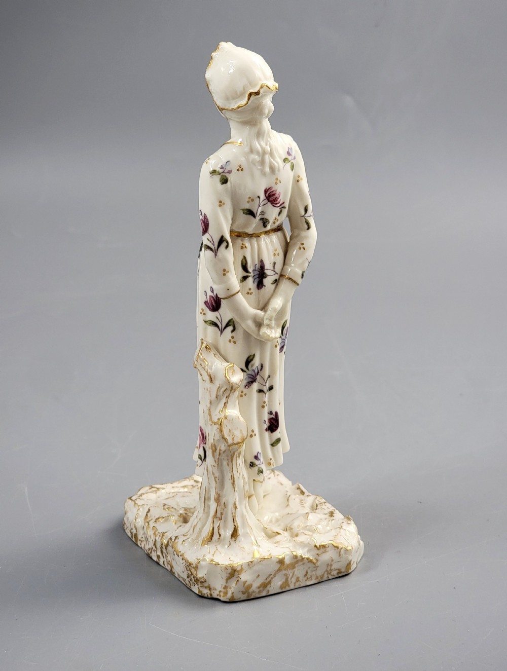 A standing porcelain figurine depicting a lady in a floral dress and bonnet with gilded trim on a textured base. View III