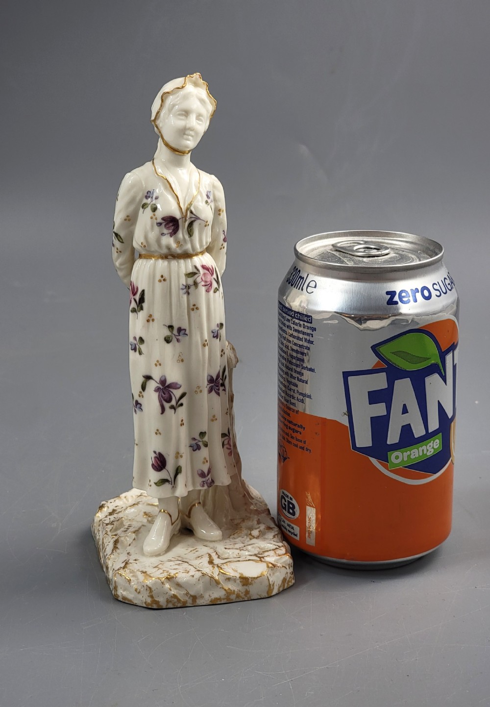 A standing porcelain figurine depicting a lady in a floral dress and bonnet with gilded trim on a textured base. View IX