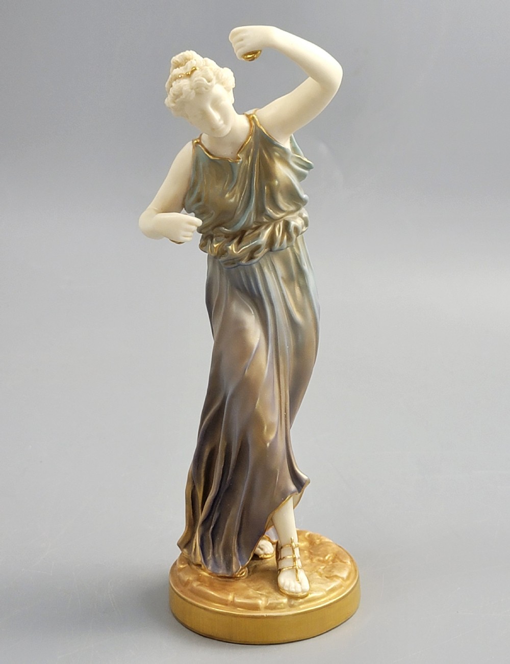 A porcelain figurine of a woman wearing classical draped robes with gilded trim standing on a circular base. View II
