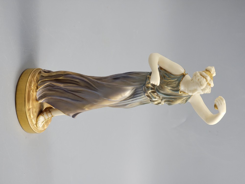A porcelain figurine of a woman wearing classical draped robes with gilded trim standing on a circular base. View III