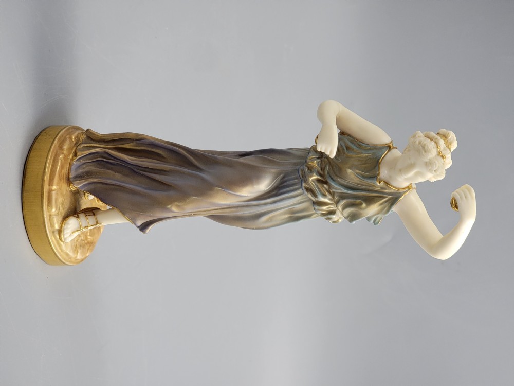 A porcelain figurine of a woman wearing classical draped robes with gilded trim standing on a circular base. View IV