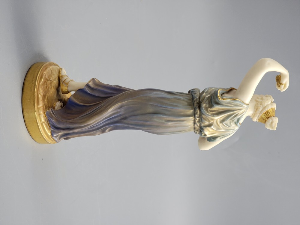 A porcelain figurine of a woman wearing classical draped robes with gilded trim standing on a circular base. View VIII