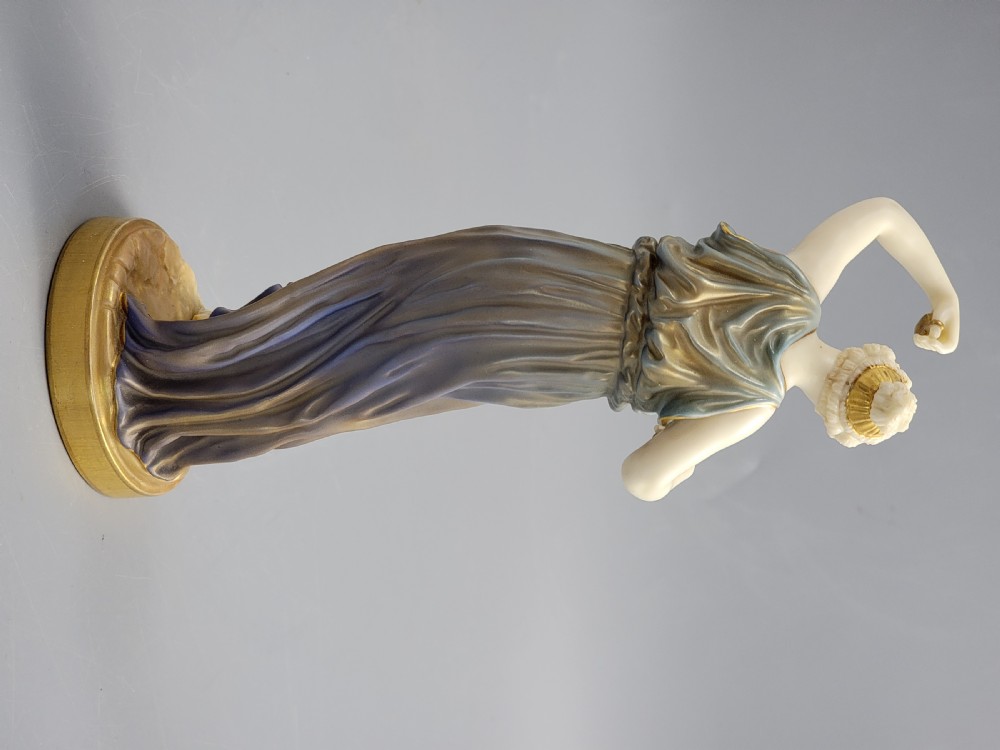 A porcelain figurine of a woman wearing classical draped robes with gilded trim standing on a circular base. View IX
