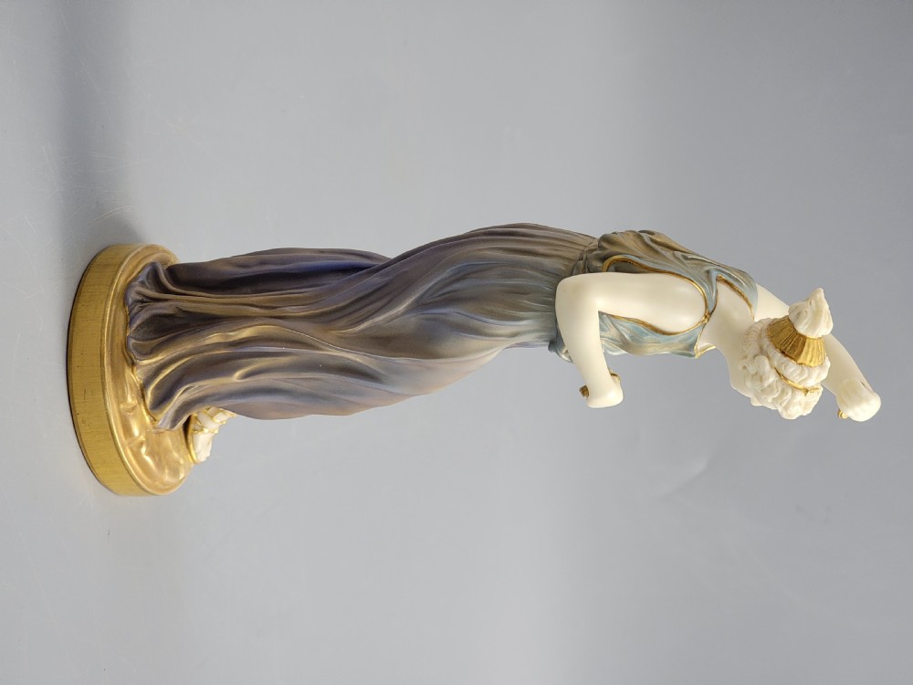 A porcelain figurine of a woman wearing classical draped robes with gilded trim standing on a circular base. View X