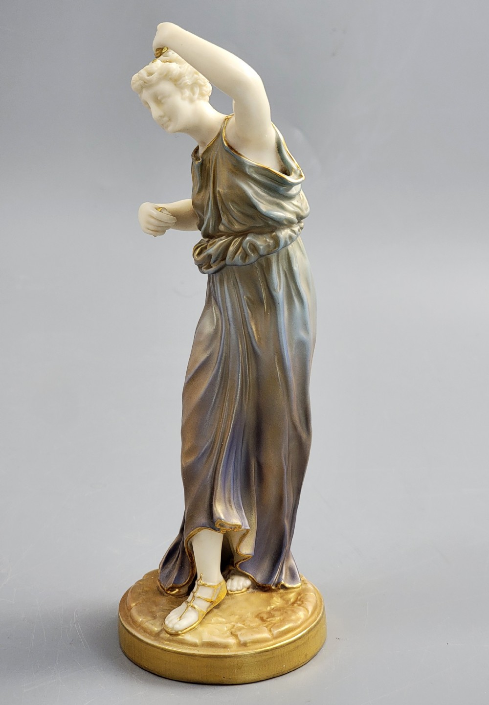 A porcelain figurine of a woman wearing classical draped robes with gilded trim standing on a circular base. View XI