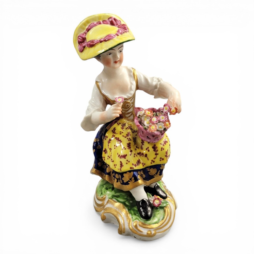 An eighteenth-century Derby porcelain figurine depicting a seated female flower seller wearing a yellow bonnet and patterned skirt while holding a small basket of flowers.