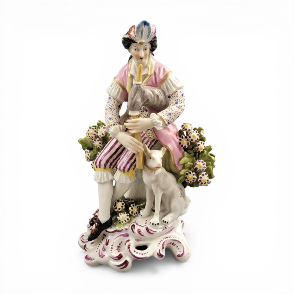 An 18th-century Derby soft-paste porcelain figurine featuring a seated bagpiper in a pink coat with a small white dog at his feet on a scrollwork base.