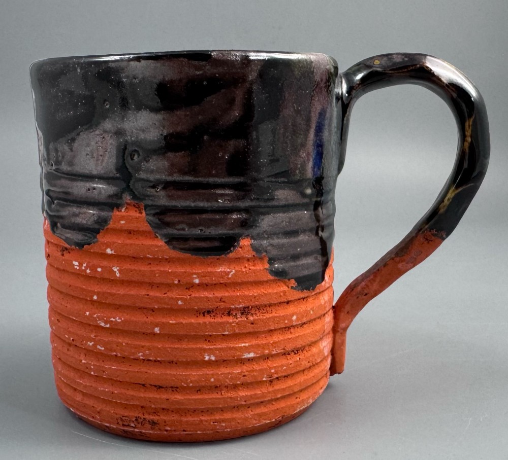 A heavy earthenware mug featuring a ribbed terracotta lower section, dark black glaze dripping from the rim, and an applied ceramic figure of a boy in a blue and white robe climbing the side. View III