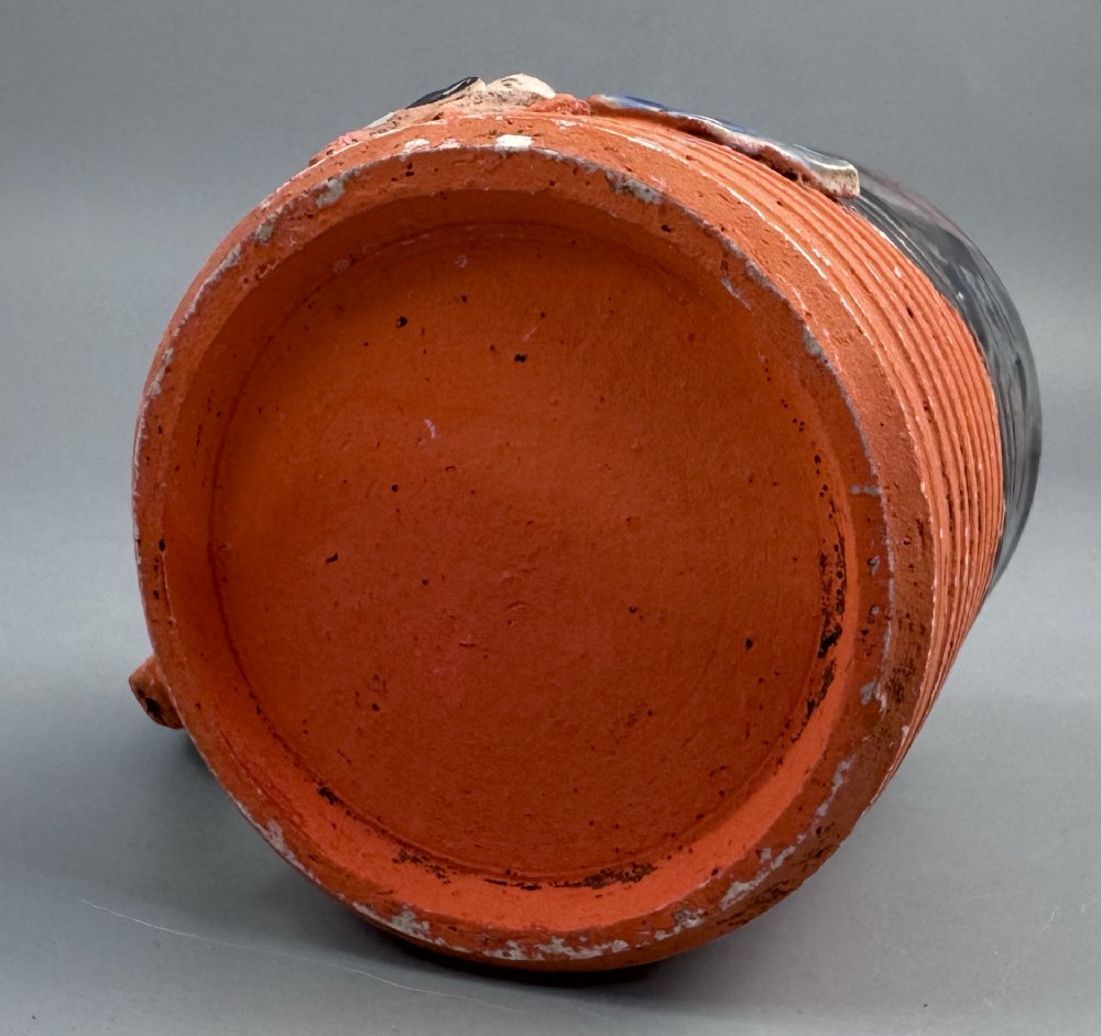 A heavy earthenware mug featuring a ribbed terracotta lower section, dark black glaze dripping from the rim, and an applied ceramic figure of a boy in a blue and white robe climbing the side. View V