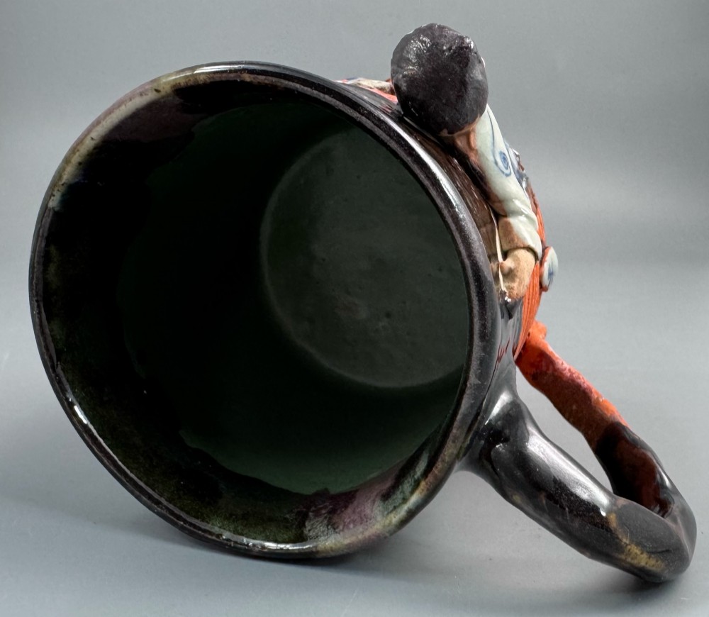A heavy earthenware mug featuring a ribbed terracotta lower section, dark black glaze dripping from the rim, and an applied ceramic figure of a boy in a blue and white robe climbing the side. View VI