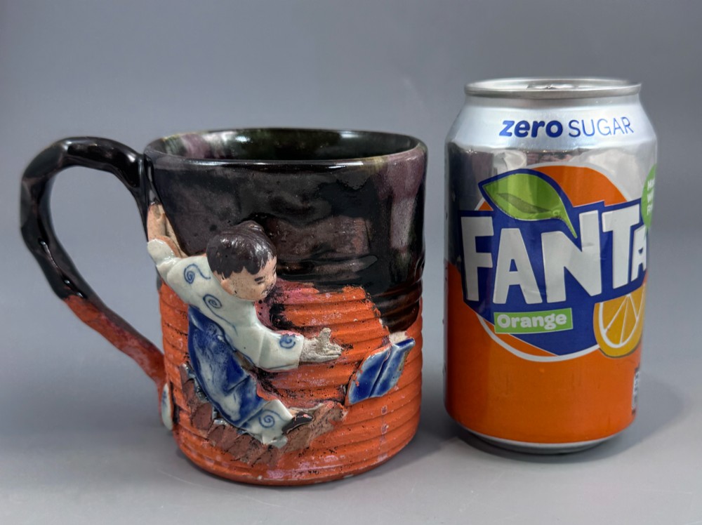 A heavy earthenware mug featuring a ribbed terracotta lower section, dark black glaze dripping from the rim, and an applied ceramic figure of a boy in a blue and white robe climbing the side. View VII