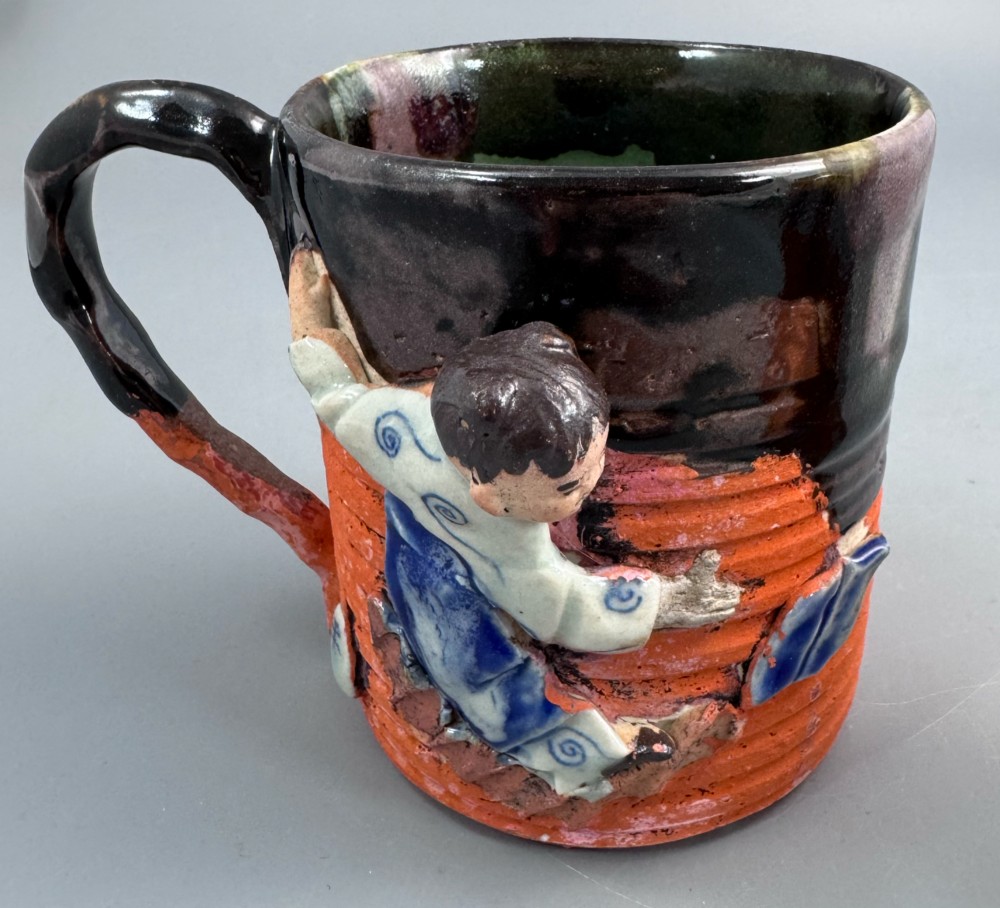 A heavy earthenware mug featuring a ribbed terracotta lower section, dark black glaze dripping from the rim, and an applied ceramic figure of a boy in a blue and white robe climbing the side. View VIII