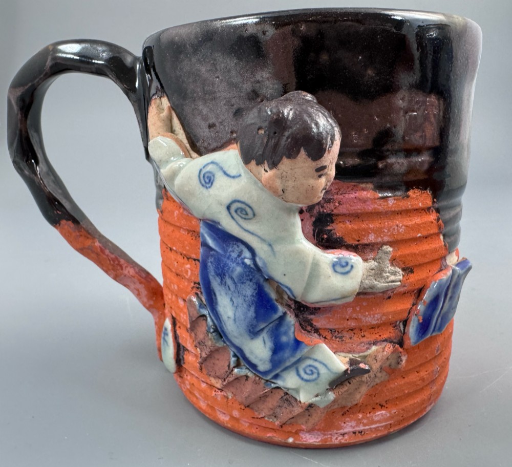 A heavy earthenware mug featuring a ribbed terracotta lower section, dark black glaze dripping from the rim, and an applied ceramic figure of a boy in a blue and white robe climbing the side. View IX