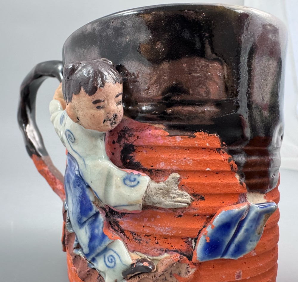 A heavy earthenware mug featuring a ribbed terracotta lower section, dark black glaze dripping from the rim, and an applied ceramic figure of a boy in a blue and white robe climbing the side. View X