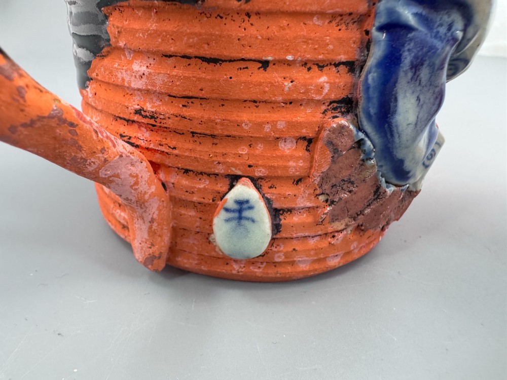 A heavy earthenware mug featuring a ribbed terracotta lower section, dark black glaze dripping from the rim, and an applied ceramic figure of a boy in a blue and white robe climbing the side. View XI