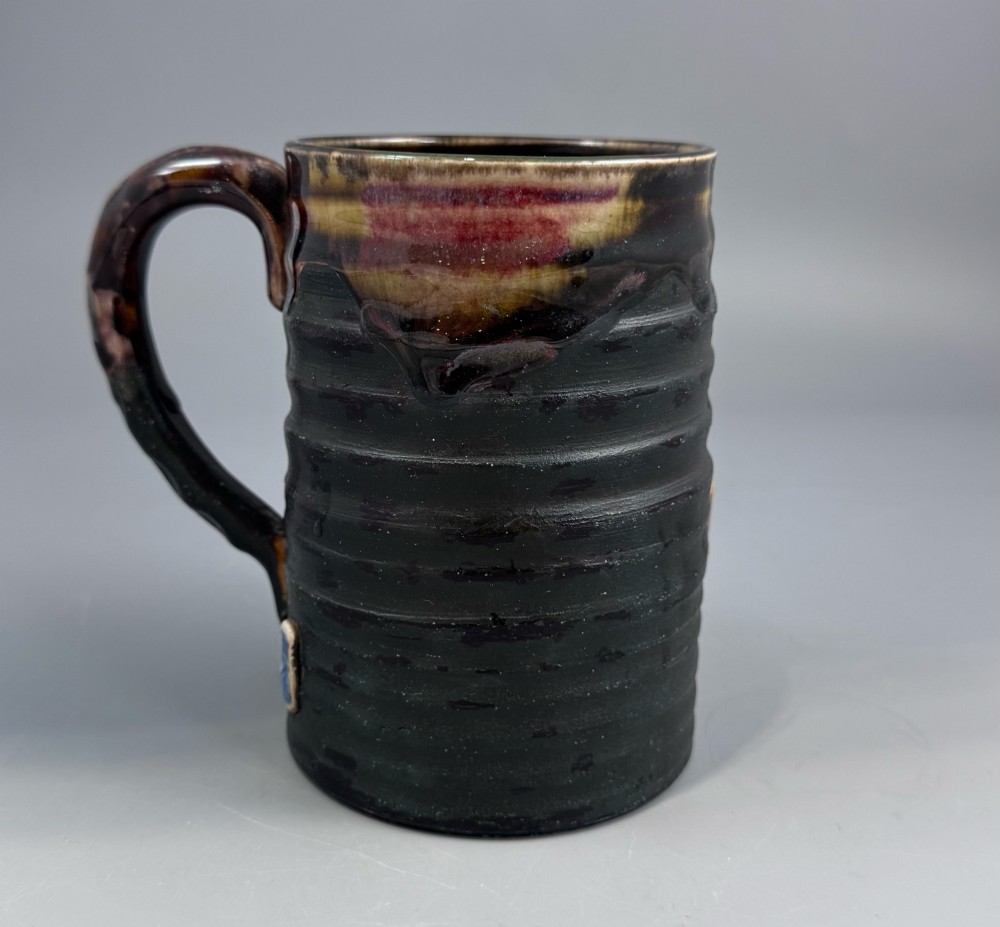 A dark ribbed ceramic mug featuring a dripping flambe glaze rim and an applied relief figure of a seated Japanese sennin. View III
