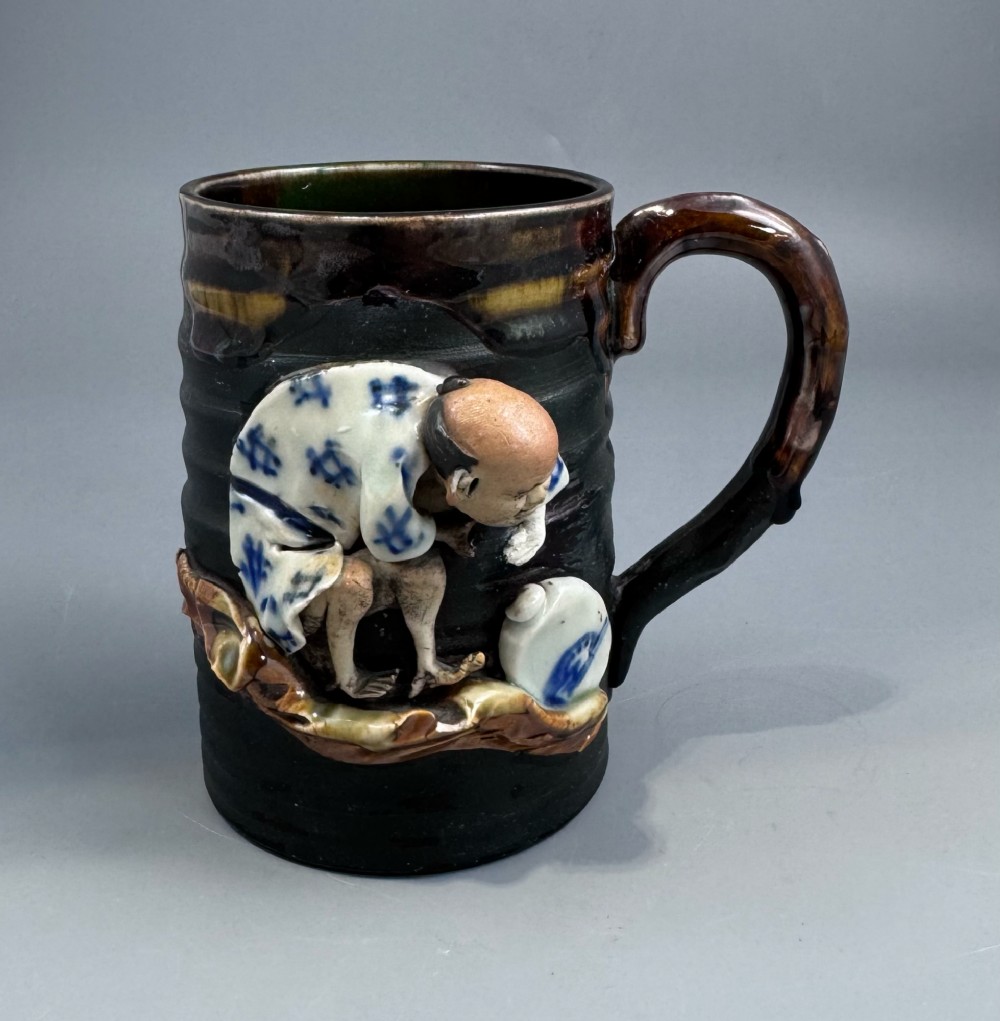 A dark ribbed ceramic mug featuring a dripping flambe glaze rim and an applied relief figure of a seated Japanese sennin.