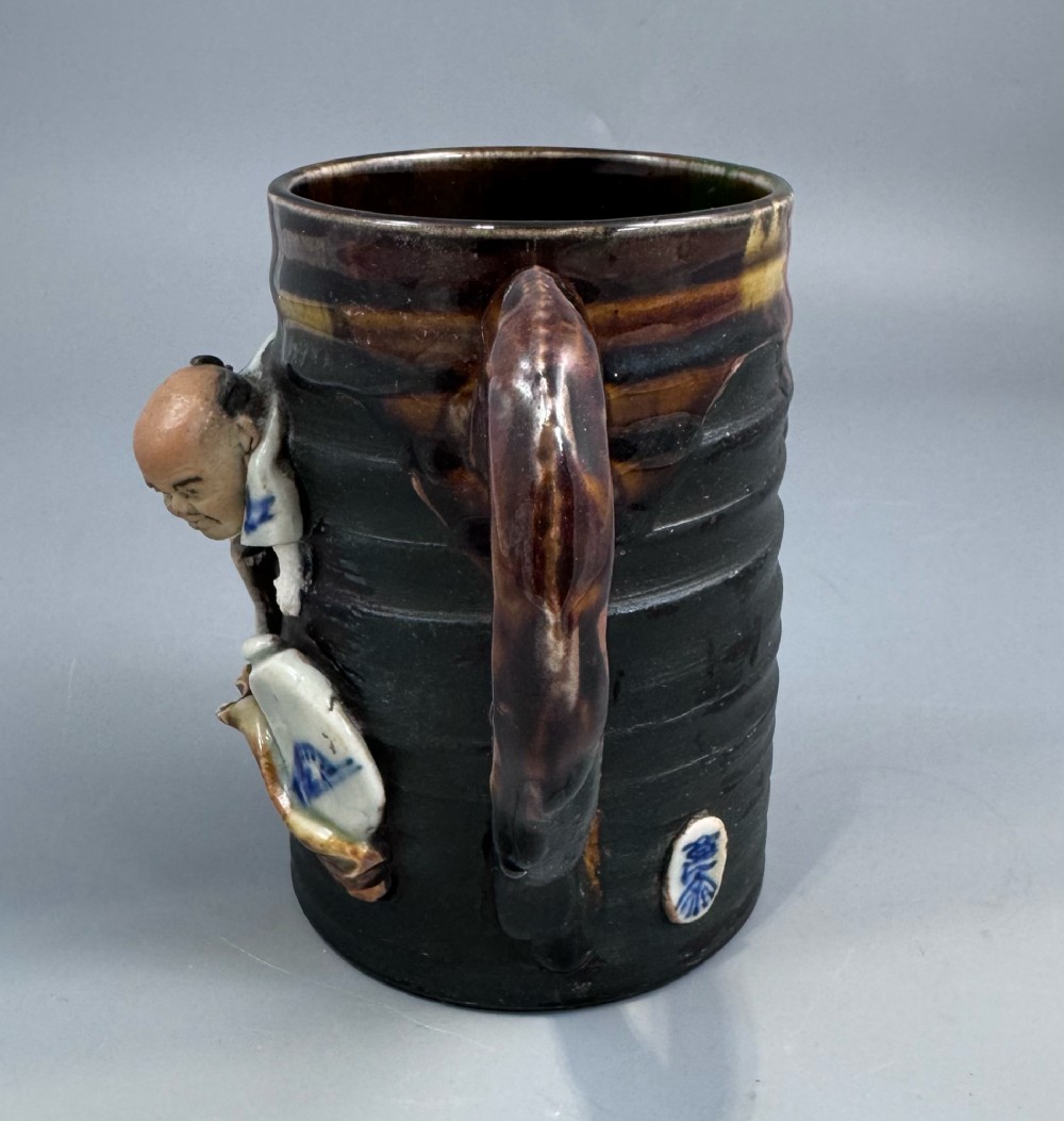 A dark ribbed ceramic mug featuring a dripping flambe glaze rim and an applied relief figure of a seated Japanese sennin. View VI