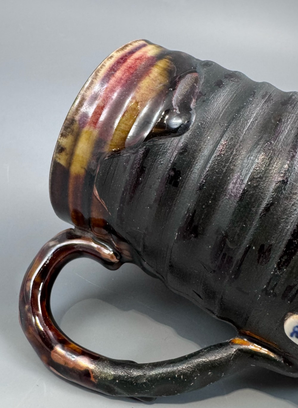 A dark ribbed ceramic mug featuring a dripping flambe glaze rim and an applied relief figure of a seated Japanese sennin. View VII