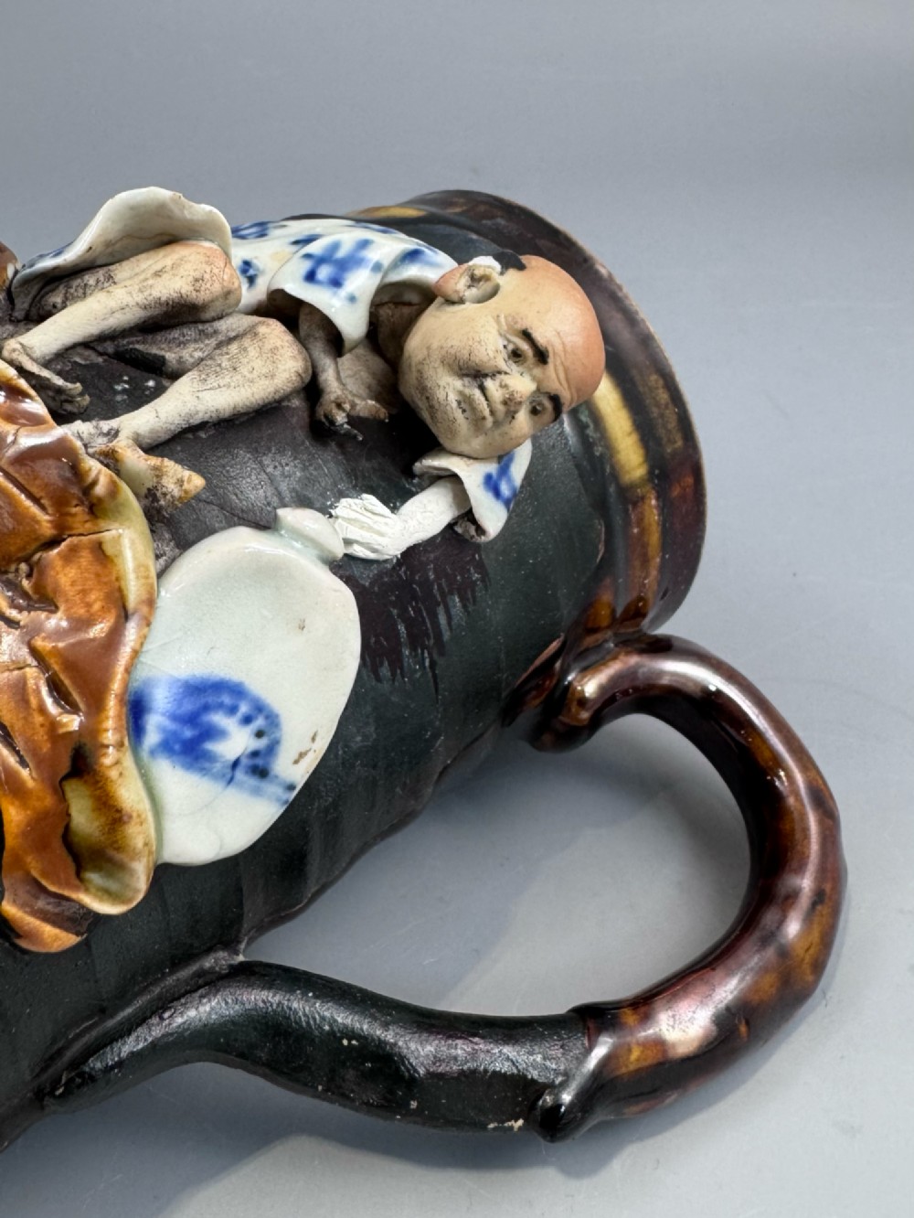 A dark ribbed ceramic mug featuring a dripping flambe glaze rim and an applied relief figure of a seated Japanese sennin. View IX